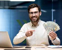 Smiling businessman siting at his desk with a laptop computer, wearing glasses and a beard, pointing at a lot of money he is holding in his right hand from using R54Media's "Local Lead Machine"